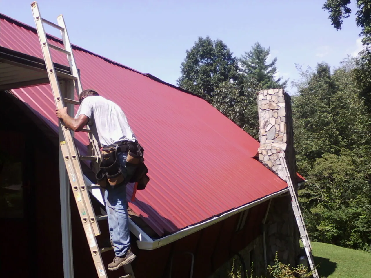Roofer on a ladder beside a red metal roof and stone chimney — Soffit & Fascia Repair by Roofing Brothers in Healdton